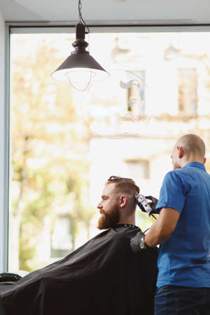 Male professional hairdresser serving client by clipper. Ginger handsome brutal stylish young man...