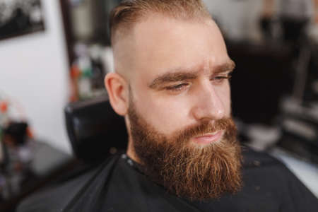 Male professional hairdresser serving client. Ginger handsome brutal stylish young man with thick...