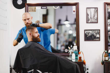 Male professional hairdresser serving client by scissors. Ginger handsome brutal stylish young man with thick big beard, short hair getting trendy haircut in black cape. Light white barber shop roomの写真素材
