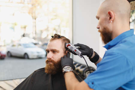Male professional hairdresser serving client by clipper. Ginger handsome brutal stylish young man...