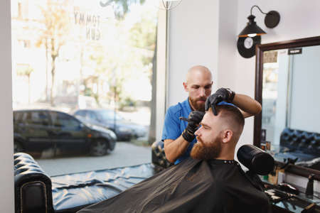 Male professional hairdresser serving client, shaving thick big beard straight razor. Ginger handsome stylish young man with short hair getting trendy haircut, black cape. Light white barber shopの写真素材