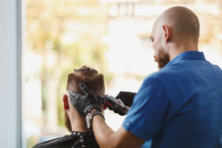 Male professional hairdresser serving client by clipper. Ginger handsome brutal stylish young man with thick big beard and short hair getting trendy haircut in black cape. Light white barber shopの写真素材