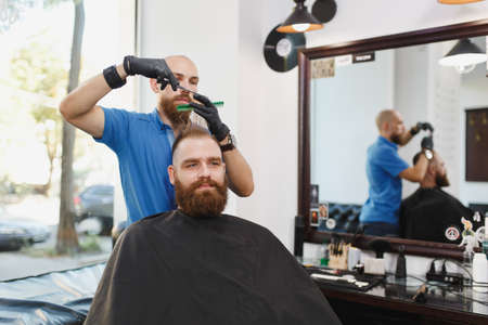 Male professional hairdresser serving client by scissors. Ginger handsome brutal stylish young man with thick big beard, short hair getting trendy haircut in black cape. Light white barber shop roomの写真素材