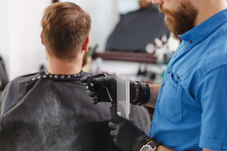 Male professional hairdresser serving and preparing tools. Ginger handsome brutal stylish young man with big beard and short hair getting trendy haircut in black cape. Light white barber shop roomの写真素材