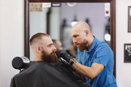 Male professional hairdresser serving client, shaving thick big beard straight razor. Ginger handsome stylish young man with short hair getting trendy haircut, black cape. Light white barber shopの写真素材