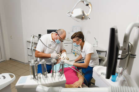 Process of preparing and installing dental ceramic crown. Male professional dentist and female assistant helps to treat the teeth of woman patient in clinic light office with modern tools equipmentの写真素材