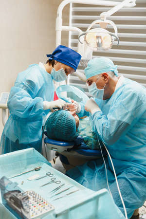 Male old professional dentist surgeon in uniform and female assistant helps to performing operation install dental implant teeth of woman patient in clinic light office with modern tools equipmentの写真素材