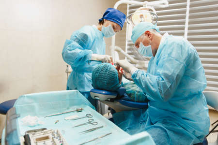 Male old professional dentist surgeon in uniform and female assistant helps to performing operation install dental implant teeth of woman patient in clinic light office with modern tools equipmentの写真素材