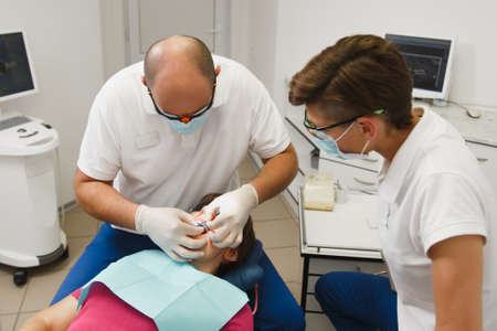 Process of preparing and installing dental ceramic crown. Male professional dentist and female assistant helps to treat the teeth of woman patient in clinic light office with modern tools equipmentの写真素材