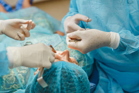 Male old professional dentist surgeon in uniform and female assistant helps to performing operation install dental implant teeth of woman patient in clinic light office with modern tools equipmentの写真素材
