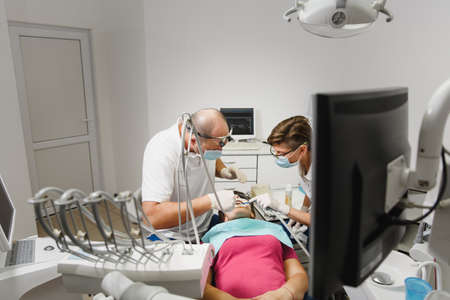 Process of preparing and installing dental ceramic crown. Male professional dentist and female assistant helps to treat the teeth of woman patient in clinic light office with modern tools equipmentの写真素材
