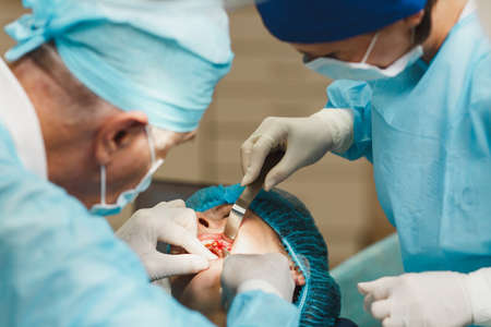 Male old professional dentist surgeon in uniform and female assistant helps to performing operation install dental implant teeth of woman patient in clinic light office with modern tools equipmentの写真素材