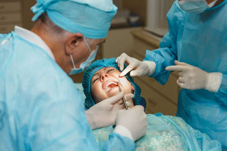 Male old professional dentist surgeon in uniform and female assistant helps to performing operation...