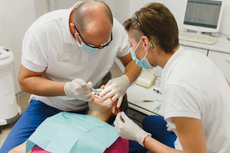 Process of preparing and installing dental ceramic crown. Male professional dentist and female assistant helps to treat the teeth of woman patient in clinic light office with modern tools equipmentの写真素材