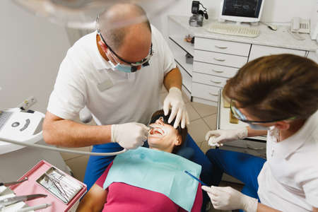 Process of preparing and installing dental ceramic crown. Male professional dentist and female assistant helps to treat the teeth of woman patient in clinic light office with modern tools equipmentの写真素材
