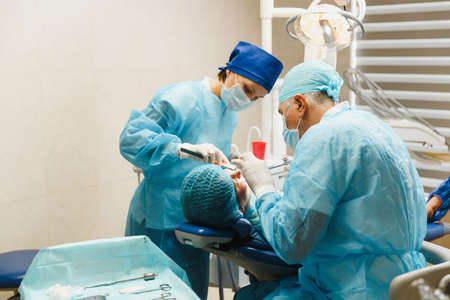 Male old professional dentist surgeon in uniform and female assistant helps to performing operation install dental implant teeth of woman patient in clinic light office with modern tools equipmentの写真素材