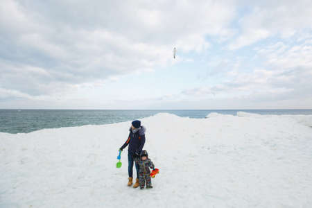 Woman play at sea snow beach background with little cute child baby boy in overalls with toy car, shovel. Mother, kid son fun on winter day. Family holiday, 15 of may, love parents, children conceptの写真素材