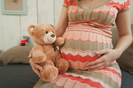 Close up cropped portrait pregnant happy woman in striped long dress with big belly, teddy bear. Tummy toy. Pregnancy, parenthood, family, motherhood, parents, children people and expectation conceptの写真素材