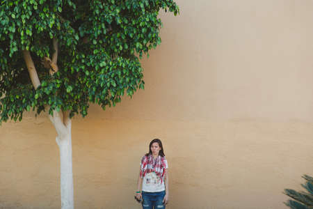 Beautiful young adorable brunette caucasian woman standing at wall of house background near green tree. Building in the cityの写真素材