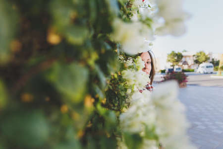 Portrait of young attractive beautiful cutie brunette woman standing in summer warm sunny day in green bushes with white flowers in front of stone rock fence. Sensuality. Side viewの写真素材