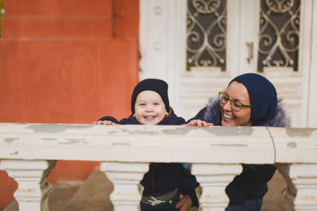Young woman with little cute child baby boy look out from behind the handrail railing with columns. Mother, little kid son have fun. Parenthood, family day 15 of may, love, parents, children conceptの写真素材