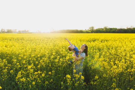 Joyful woman walk on green yellow flowering field background, rest, have fun, play, toss up little cute child baby boy. Mother, little kid son. Family day 15 of may, love, parents, children conceptの写真素材