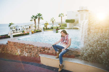 Beautiful young brunette caucasian woman sitting on poolside in jeans, handkerchief, t-shirt, yellow boots relaxing with waterfall and sea in background. Concept of tourism, vacation, recreationの写真素材