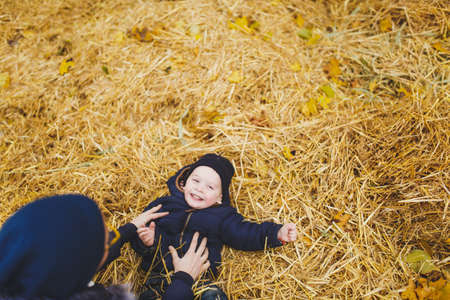 Little cute child baby boy walk in fall park, sit, lay in pile of straw, have fun in dry yellow autumn grass. Mother fun tickle kid son. Parenthood family day 15 of may love parents, children conceptの写真素材