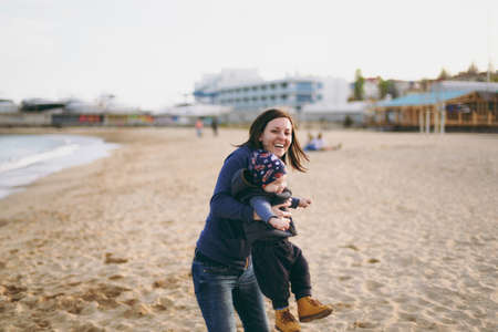 Young woman playing at sea sand beach background with little cute child baby boy, good mood. Mother, little kid son on sunny day. Parenthood, family holiday 15 of may, love, parents, children conceptの写真素材