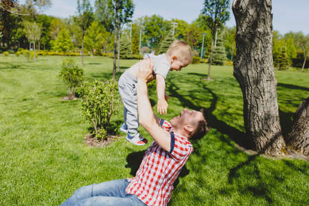 Joyful man walk on green park, rest, have fun, play, high toss up little cute child baby boy sit on grass at tree. Father throw up little kid son. Family day 15 of may, love parents, children conceptの写真素材