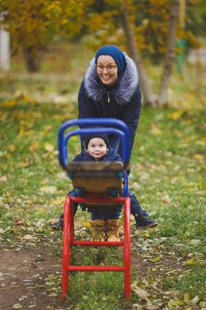 Young woman walk in green yellow fall autumn park with little cute child baby boy on nature. Mother, kid son fun rest, ride on swing. Parenthood, family day 15 of may, love, parents, children conceptの写真素材
