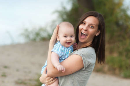 Young fun smiling woman standing at sea background hugging little cute child baby boy on nature, green trees. Mother, little kid son. Parenthood, family day 15 of may, love, parents, children conceptの写真素材