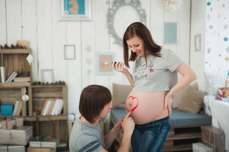 Pregnant woman with big belly. man paints heart with paints on tummy. Couple with kid clothes. Wife, husband. Pregnancy, parenthood, family, motherhood, parents, children, people, expectation conceptの写真素材
