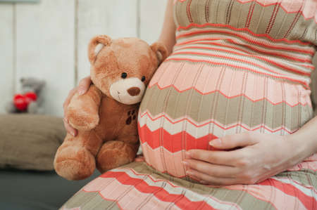 Close up cropped portrait pregnant happy woman in striped long dress with big belly, teddy bear. Tummy toy. Pregnancy, parenthood, family, motherhood, parents, children people and expectation conceptの写真素材