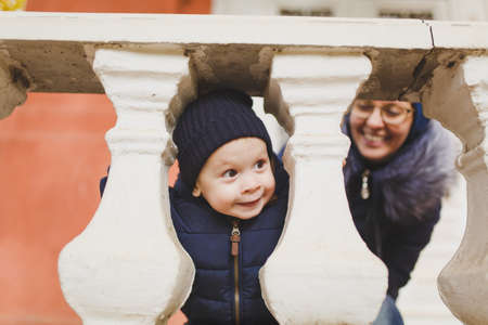 Young woman with little cute child baby boy look out from behind the handrail railing with columns. Mother, little kid son have fun. Parenthood, family day 15 of may, love, parents, children conceptの写真素材