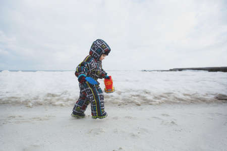 Little cute child baby boy in overalls play at sea cover snow white beach background with toy car, shovel. Small kid son fun on winter day. Family holiday, 15 of may, love, parents, children conceptの写真素材