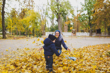 Young woman walk in fall park with little cute child baby boy, toss throw up dry yellow autumn leaves. Mother, little kid son rest. Parenthood, family day 15 of may, love, parents, children conceptの写真素材