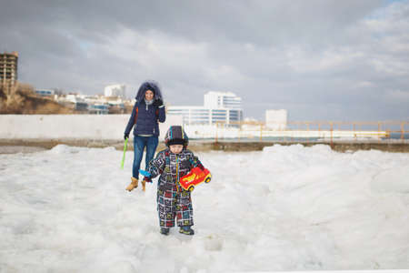 Woman play at sea snow beach background with little cute child baby boy in overalls with toy car, shovel. Mother, kid son fun on winter day. Family holiday, 15 of may, love parents, children conceptの写真素材