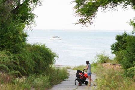 Young woman walking at sea background with little cute child baby boy in baby carriage on nature, green trees. Mother, little kid son. Parenthood, family day 15 of may, love, parents children conceptの写真素材