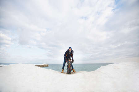 Woman play at sea snow beach background with little cute child baby boy in overalls with toy car, shovel. Mother, kid son fun on winter day. Family holiday, 15 of may, love parents, children conceptの写真素材