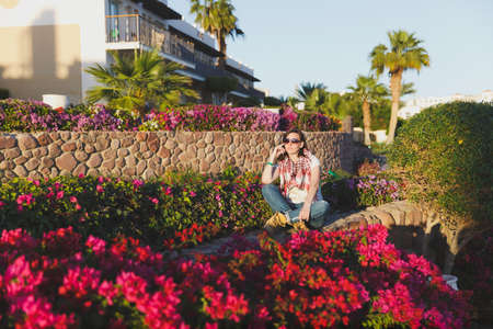Portrait of young attractive beautiful cutie brunette woman standing in summer warm sunny day in green bushes with pink flowers in front of stone rock fence. Sensualityの写真素材