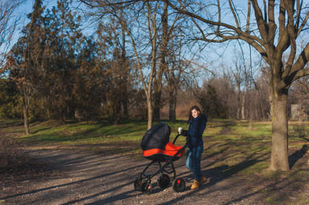 Young woman in blue coat walking in autumn park with little child baby in orange baby carriage, tea or coffe on fall nature, green trees. Mother and kid. Parenthood, love, parents, children conceptの写真素材