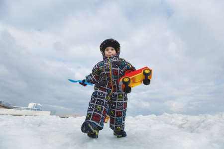 Little cute child baby boy in overalls play at sea cover snow white beach background with toy car,...