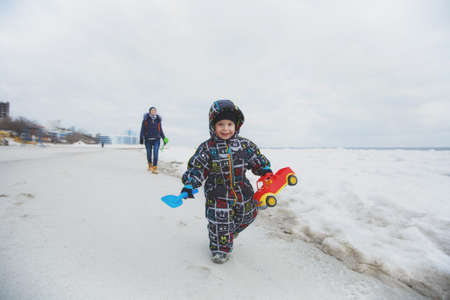 Woman play at sea snow beach background with little cute child baby boy in overalls with toy car, shovel. Mother, kid son fun on winter day. Family holiday, 15 of may, love parents, children conceptの写真素材