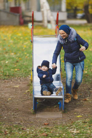 Young woman walk in green yellow fall autumn park with little cute child baby boy on nature. Mother, kid son fun rest, ride children s slides on playground. Family day 15 of may, love parents conceptの写真素材