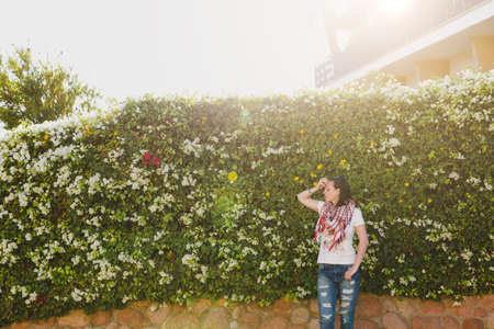 Portrait of young attractive beautiful cutie brunette woman standing in summer warm sunny day in green bushes with white flowers in front of stone rock fence.の写真素材