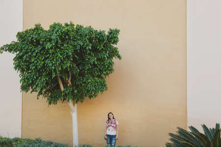 Beautiful young adorable brunette caucasian woman standing at wall of house background near green tree. Building in the cityの写真素材