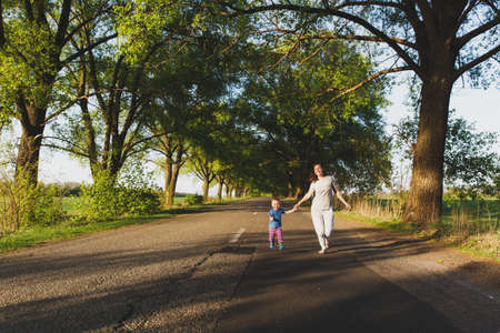 Young woman, little cute child boy walk on road with trees on the roadside, catch up, run in warm summer day. Mother, kid son rest. Parenthood, family day, 15 of may, love, parents, children conceptの写真素材