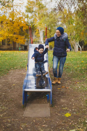 Young woman walk in green yellow fall autumn park with little cute child baby boy on nature. Mother, kid son fun rest, ride children s slides on playground. Family day 15 of may, love parents conceptの写真素材