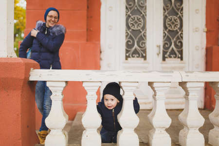Young woman with little cute child baby boy look out from behind the handrail railing with columns. Mother, little kid son have fun. Parenthood, family day 15 of may, love, parents, children conceptの写真素材
